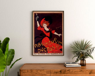 Póster publicitario vintage Absinthe Ducros, enmarcado sobre una cómoda de madera, entre plantas.