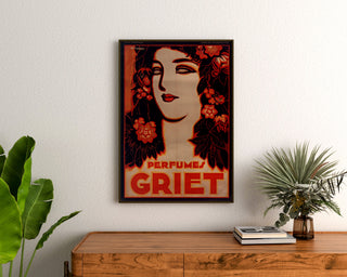Póster publicitario vintage Perfumes Griet, enmarcado sobre una cómoda de madera, entre plantas.