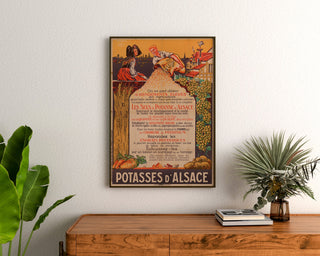 Póster publicitario vintage Potasses d’Alsace, enmarcado sobre una cómoda de madera, entre plantas.