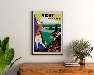 Póster turístico vintage – Vichy, manantiales y turismo, enmarcado sobre una cómoda de madera, entre plantas.