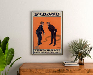 Cartel vintage A Brace of Partridges, Strand Theatre, enmarcado sobre una cómoda de madera, entre plantas.
