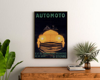 Póster vintage Automoto, Saint‑Étienne, enmarcado sobre una cómoda de madera entre plantas.