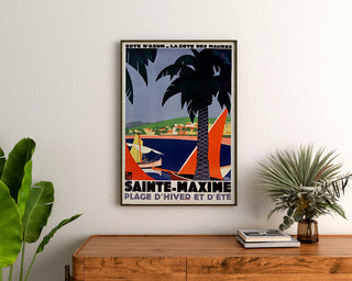 Póster vintage Sainte-Maxime, costa de los Maures, enmarcado sobre una cómoda de madera, entre plantas.