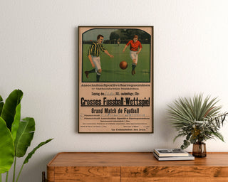 Póster vintage Sarreguemines, partido de fútbol, enmarcado sobre una cómoda de madera, entre plantas.